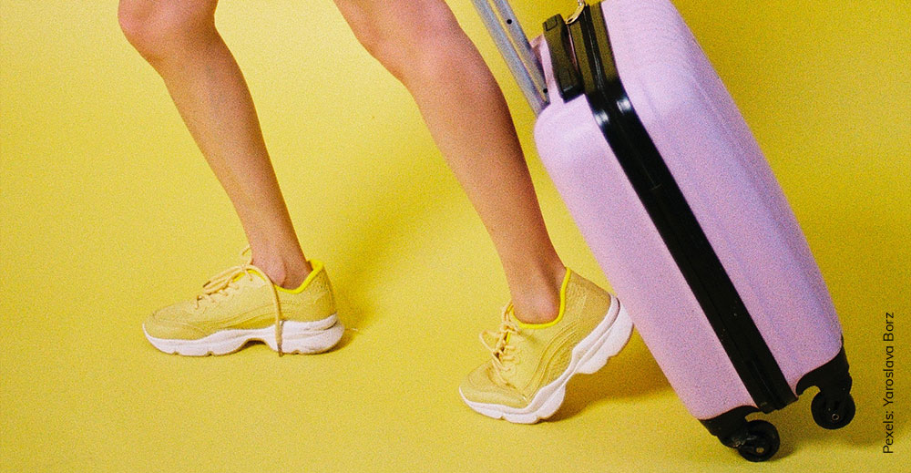 Beine einer weiblichen Person in Turnschuhen die einen lila Koffer hinter sich her zieht